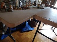 Top side view of a folding table showing beige plastic top and corner edge with black metal folding legs beneath.