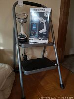 Folding 2-step stool with black anti-slip bottom tread and missing top tread alongside Rowenta handheld steamer on top step