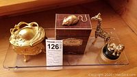 Four items on clear display tray: brass bombe chest style keepsake box, miniature box with MGM dusting powder, brass giraffe figurine, brass mouse figurine