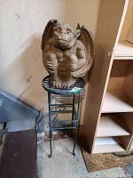 Concrete gargoyle statue sitting on green metal plant stand with four legs against wall and wood shelf.