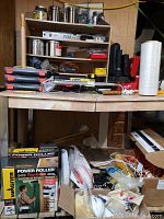 Photo showing table with Wagner Power Roller box, paint rollers, various tools, coveralls, and other hardware items under table and on shelves