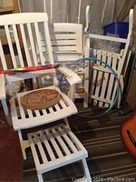 Photo showing white resin patio chairs including folding chairs and rocking chair, with umbrella attached to chair arm, hula hoop leaning on chairs, and wooden carved plaque on chair seat.