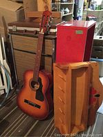 Shows guitar leaning against wooden shelf and red cabinet, with attache case on top of cabinet and part of red shelf visible