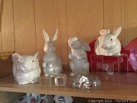 Set of four white Fitz and Floyd bunny figurines displayed alongside two glass heart paperweights and two clear acrylic risers on wooden shelf