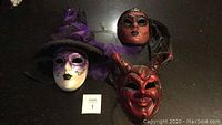 Three original handmade Italian masks: a white witch mask with purple hat and designs, a red devil mask with horns, and a brown mask with black cloth covering the back