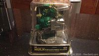 Hulk collector clock displayed inside clear protective case with metallic base, showing Hulk figure and clock mechanism.