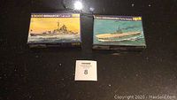 Two Heller model ship kits in box, Bismarck (1/2000 scale) and Arromanches (1/1600 scale) on black surface with lot number card.