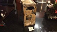 Photo of the original packaging box for a Rudolf Christmas tree watering system showing the product name and some illustrations.