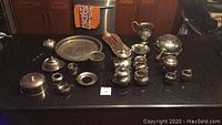 Front view of all silver plate items on black surface showing two tea sets, platters, and various serving items.