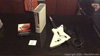 Xbox 360 console, Gibson Xbox Guitar Hero controller, and boxed PS3 component cable displayed on black surface.