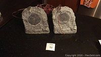 Front view of two gray rock speakers with wires on dark surface; one speaker shows internal damage and debris.
