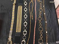 Lot of 7 assorted fashion belts laid out on black fabric, showing different metal chain styles and decorative elements.