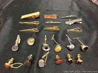 Top view of 10 tie clips and 11 decorative tie tacks arranged on black fabric.