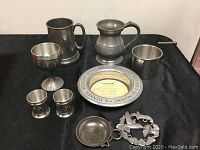 Full set laid on black cloth showing all eight pewter items including tankards, goblet, shot glasses, decorative dish, small dish and bottle stand.