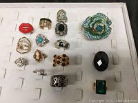 Front layout view of 15 assorted costume jewellery rings in a white display tray, showing variety of styles and colors.