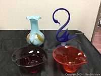Four art glass pieces displayed on black cloth: pale blue ruffled edge vase with leaf pattern, cobalt blue swan figurine, two glass bowls (amber and amethyst).
