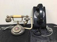 Two vintage rotary dial telephones displayed on a black surface, one black wall-mount style and one silver French-style desk phone.