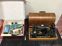 Photo showing the vintage Singer sewing machine with wooden base and roll-top wooden cover beside a box of assorted attachments and accessories.