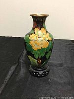 Front view of cloisonne vase showing large yellow flower and green leaves on black background with wooden stand.