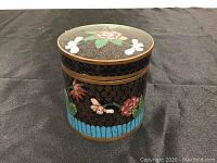 Front and side view of the round cloisonné box showing floral and butterfly enamel designs on black background with gold swirls and blue border.
