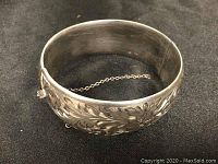 Front angle of a wide sterling silver bangle bracelet showing floral engravings and attached safety chain.