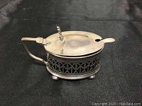 Side view of sterling silver mustard pot with hinged lid and handle on a black background.