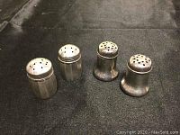 Two pairs of marked sterling silver shakers, viewed from a slightly elevated angle on a dark cloth surface, showing their overall shape and condition.