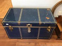 Blue metal trunk with gold-tone riveted metal strips and visible scratches on top.