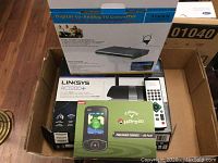Box containing the three main items: Tivax digital to analog TV converter, Linksys AC1200+ router with remote, and Calloway UproGo device with preloaded courses SD card.