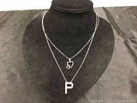 Two silver necklaces on black display. One has a double heart pendant and the other a letter "P" pendant with stones.