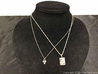 Two silver necklaces displayed on black jewelry bust; one with small trio bead pendant, one with book locket pendant with red stones