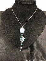 Front view of the necklace on a black display showing the silver chain and the oval turquoise pendant with dangling turquoise beads.