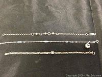Three silver bracelets laid out on black fabric: one with rings and spherical beads, one thin chain, one with geometric pattern and decorative stone.