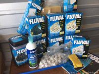 Various Fluval aquarium filter media boxes stacked together including carbon, biomax, and pre-filter sponge media with a bottle of Top Fin bacterial supplement and loose ceramic rings on table.