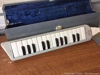 Full length view of the Hero melodica keyboard resting on a wooden surface with the protective case open behind it.