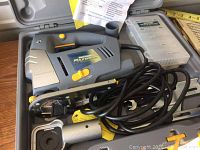 Mastercraft Maximum serpentine saw packed in hard plastic carrying case with coiled power cord, accessory box containing several saw blades, instruction manual placed over saw, and additional handle attachment stored in case.