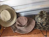 Three Tilley hats lined up on a wooden surface against a metal wall - two solid color wide-brim hats (tan and light brown) and one black patterned hat