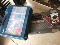 Blue Rubbermaid toolbox with transparent top compartment showing various small hardware items inside, and a black plastic case with a digital multimeter voltage tester and red/black test leads beside it.
