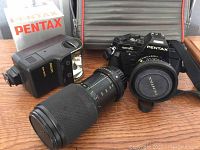 Front view of Pentax Super A camera body with 50mm lens attached, telephoto lens and AF200 flash unit positioned in front of soft carrying case and 50mm lens box.