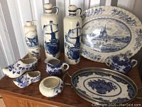 Full view of the assorted Delft blue and white china pieces showing tall bottles with windmill design, flower decorated shoe-shaped containers, small pitchers, large platter with scenic village and windmill, and oval serving tray with floral bouquet.