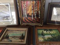 Five framed pieces of art shown on table including an oil of birch trees, a winter landscape oil painting, and a tropical scene print signed M. Endo 2010, and two other artworks partially visible.