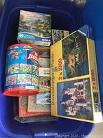 Photo showing blue bin filled with assorted jigsaw puzzle boxes including Thomas Kinkade 1000-piece puzzle and Ravensburger 2x1000-piece puzzle set