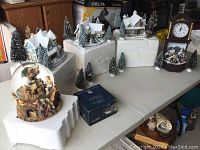 Image showing multiple Thomas Kinkade snow-covered village house sculptures and pine trees arranged on white blocks for display and storage.