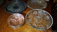 Overview showing all four items on wood table: two clear glass starburst platters, one silver engraved platter, and one rose glass divided nut bowl in silver holder.