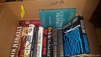 Box with hardcover books including Stephen King, James Patterson, James Redfield, and others, spines facing up.