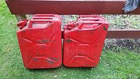 Front view of two red metal jerry cans placed side by side on green grass with some paint wear and rust showing.