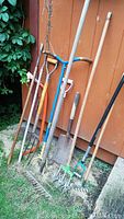 Photo showing a collection of eight assorted garden tools leaning against an orange wooden wall, including rakes, shovels, pitchforks, and cultivators with wooden and metal handles.