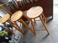 Three solid wood round seat bar stools in light wood finish, visible wear and positioned on concrete floor in front of wooden background.