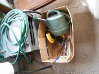 Suncast hose reel with attached green garden hose and a cardboard box containing a green watering can and various gardening tools on the floor.