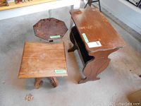 Set of three small wooden tables on concrete floor showing aged wear and stains: an end table with curvy side panels and shelving, an octagon-shaped fern stand, and a square plant stand with curved legs.